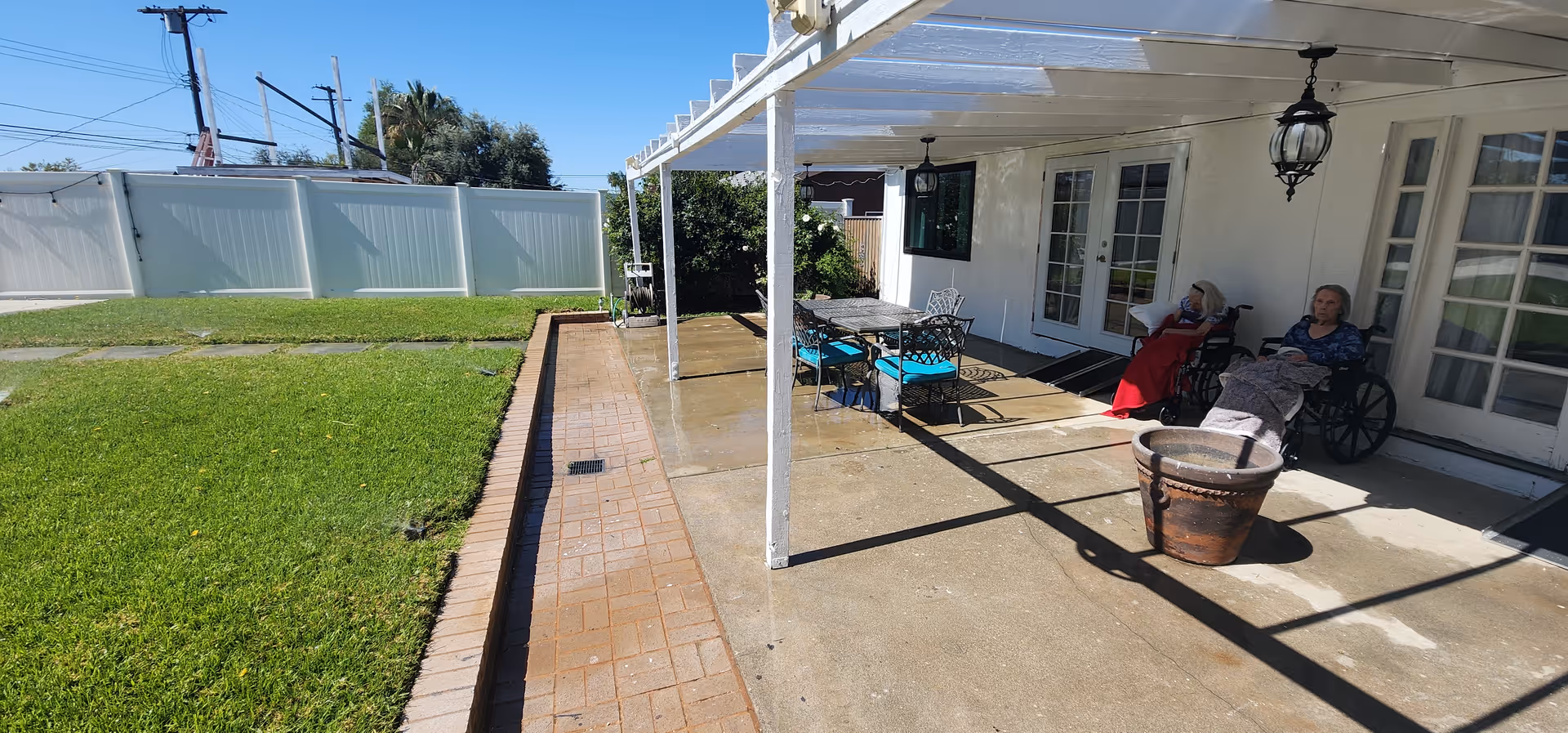 Covered patio area at Affordable Board and Care showing two residents in wheelchairs seated by a table and chairs next to a lawn.