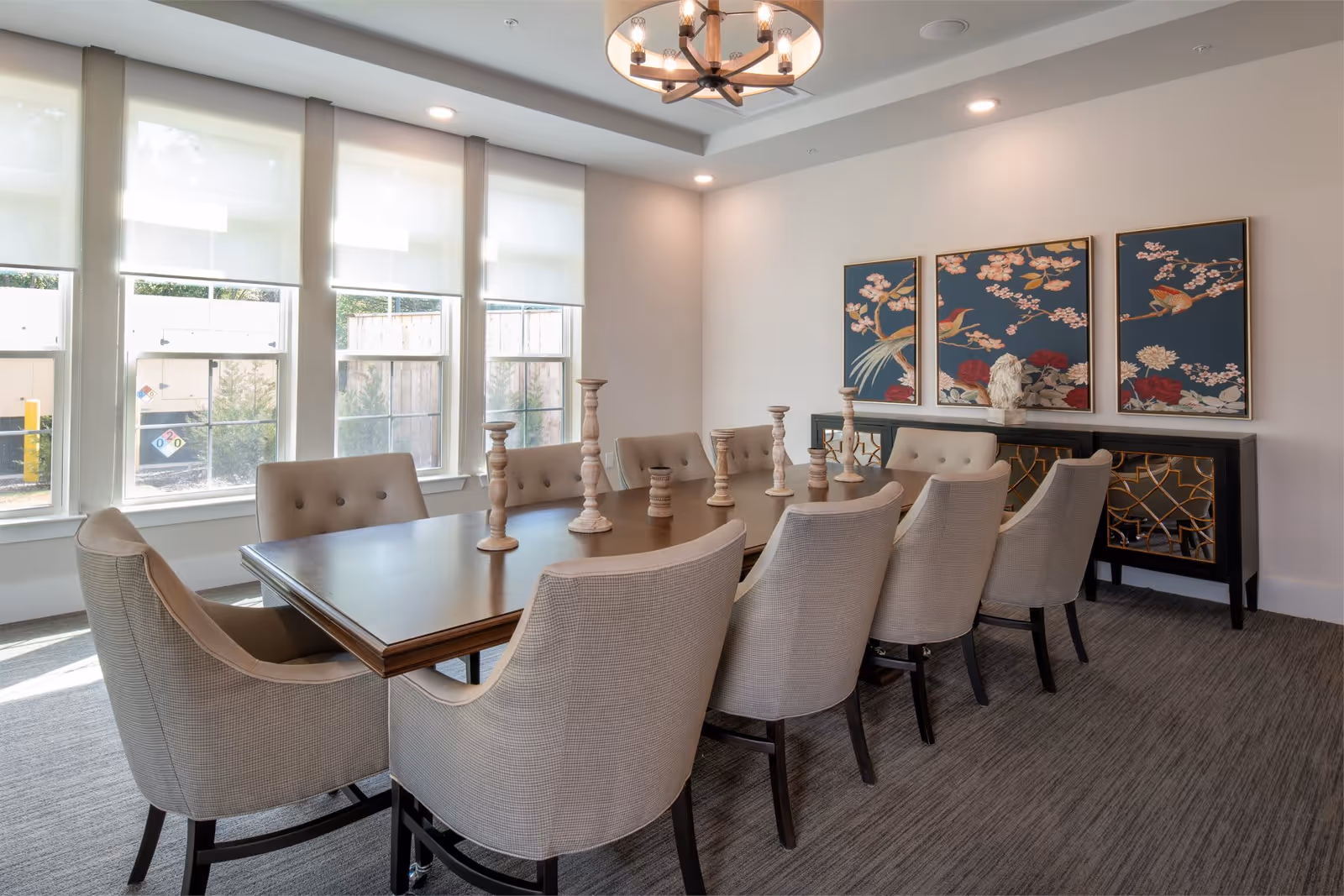 A bright dining room with a long wooden table surrounded by ten beige upholstered chairs. The table is decorated with multiple wooden candle holders. Large windows with white roller shades let in natural light. On the wall behind the table, there are three framed artworks featuring birds and flowers, and below them is a black sideboard with gold decorative accents. A modern chandelier hangs from the ceiling.