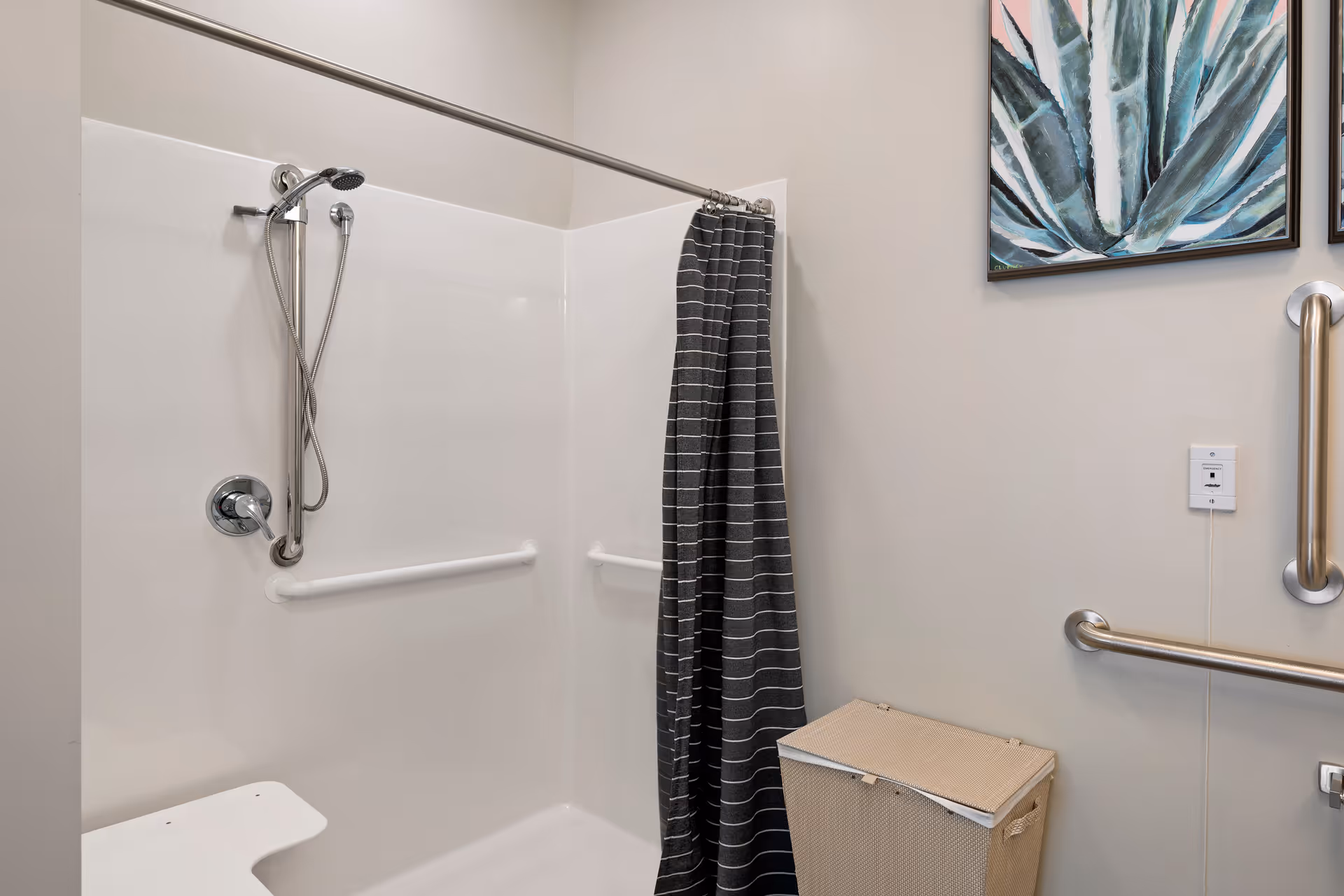 A bathroom shower area with white walls and grab bars installed for safety. There is a handheld showerhead attached to a vertical bar, a dark gray shower curtain with white stripes, a beige laundry hamper, and a pull cord emergency alarm on the wall. Two framed botanical paintings are partially visible on the wall.