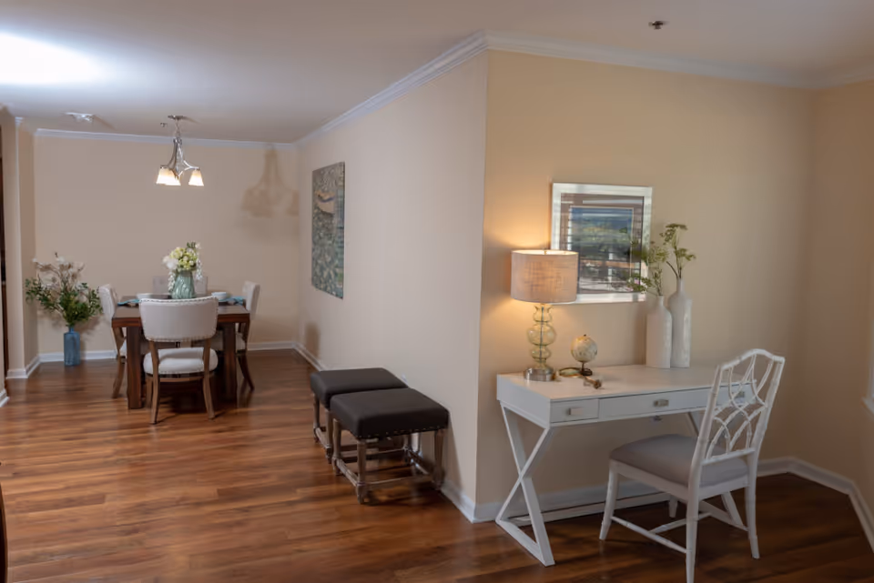 Light-filled interior showing a dining table with chairs and a small white desk with a chair and lamp against a beige wall.