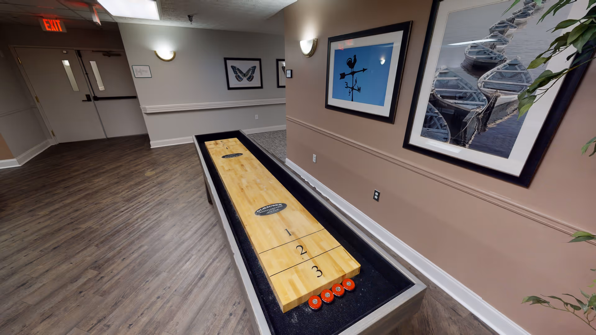 Indoor shuffleboard table in a hallway with wood flooring and beige walls. The hallway features framed artwork on the walls, including a butterfly and a weather vane. There is an exit door at the end of the hallway.