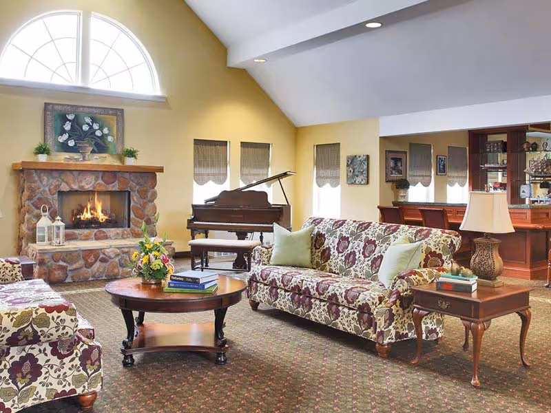 A cozy living room with floral patterned sofas, a round wooden coffee table with books and a flower arrangement, a stone fireplace with a fire burning, a grand piano near windows with blinds, and a wooden side table with a lamp and books. The room has a high ceiling with recessed lighting and a large arched window above the fireplace.
