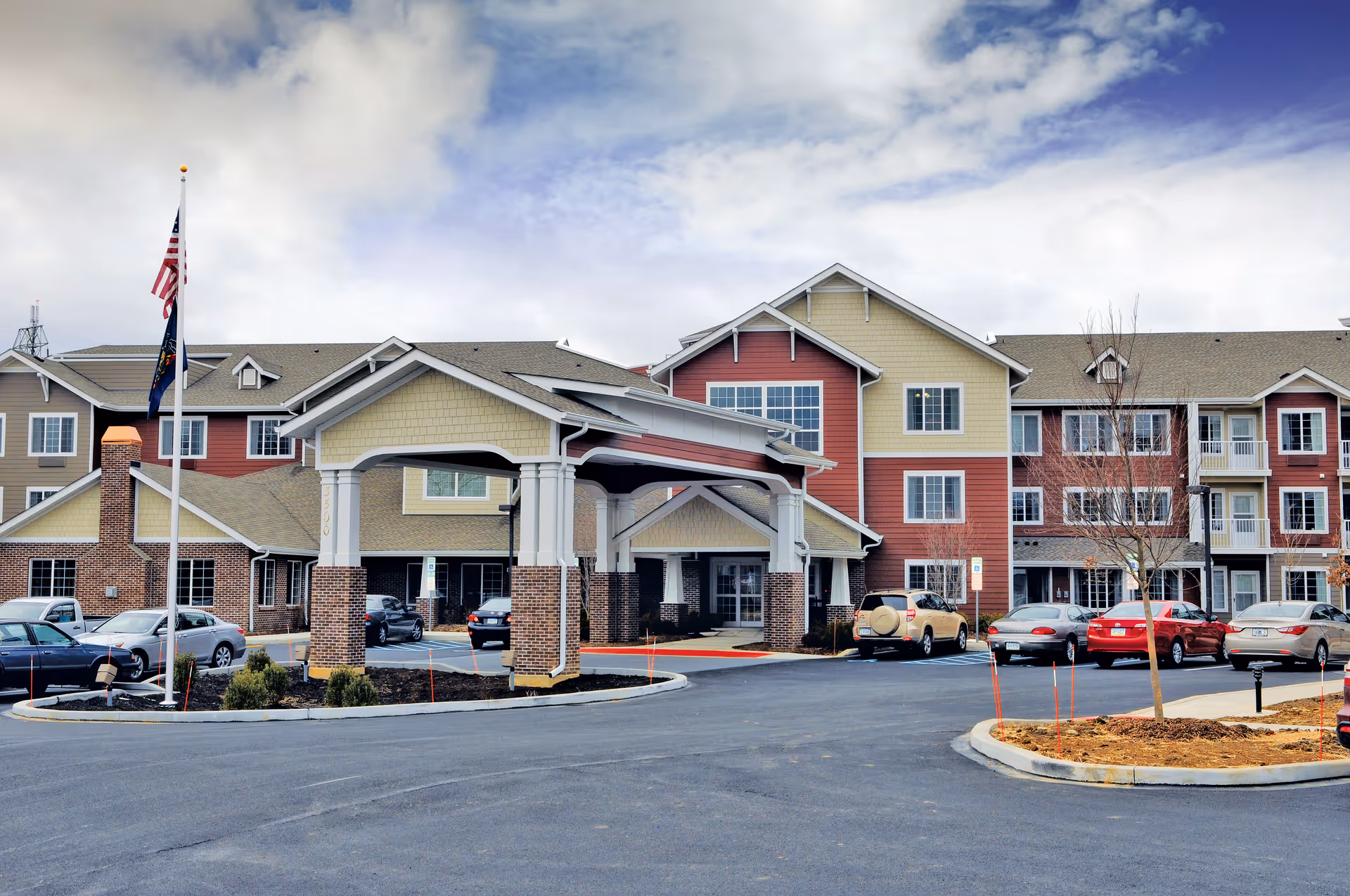 Exterior view of Sterling Heights Gracious Retirement Living facility showing a multi-story building with a covered entrance, several parked cars, and two flagpoles with flags in front under a partly cloudy sky.