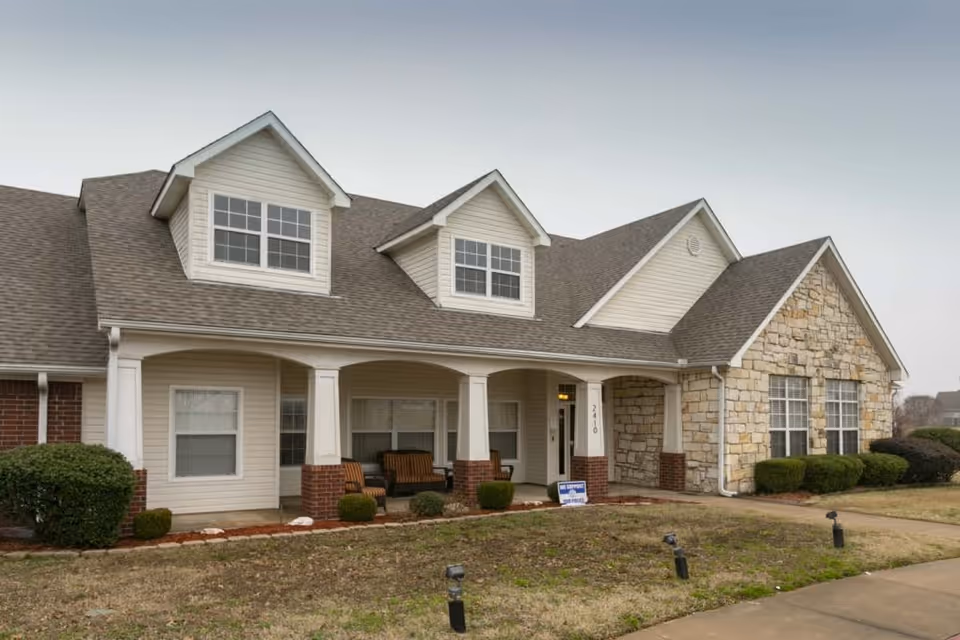 Exterior view of a single-story senior living facility building with beige siding, stone accents, and a covered porch with seating. The lawn is partially grass-covered with small bushes and a paved walkway leading to the entrance.