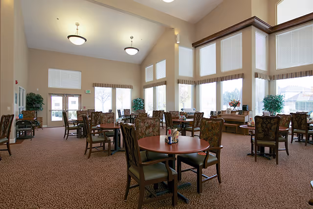 Spacious dining room with round wooden tables and upholstered chairs beneath high ceilings and large windows.