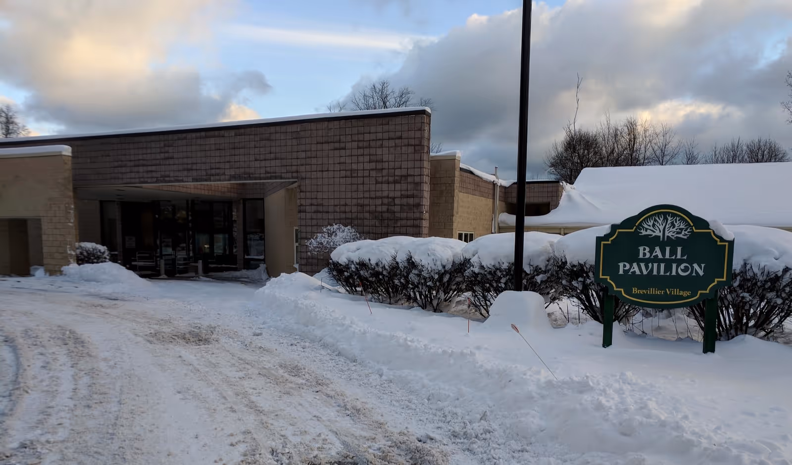 Snow-covered driveway and entrance to a low brick building with a green sign reading 'Ball Pavilion Brevillier Village'.
