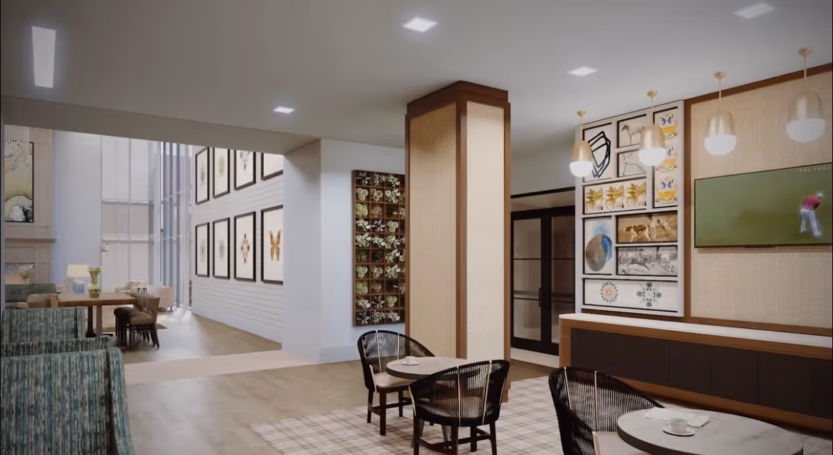 A modern and spacious interior common area with seating arrangements including tables and chairs. The room features a wall-mounted TV showing a golf game, decorative framed artwork on the walls, and pendant lights hanging from the ceiling. There is a mix of comfortable chairs and a dining table visible in the background near large windows letting in natural light.