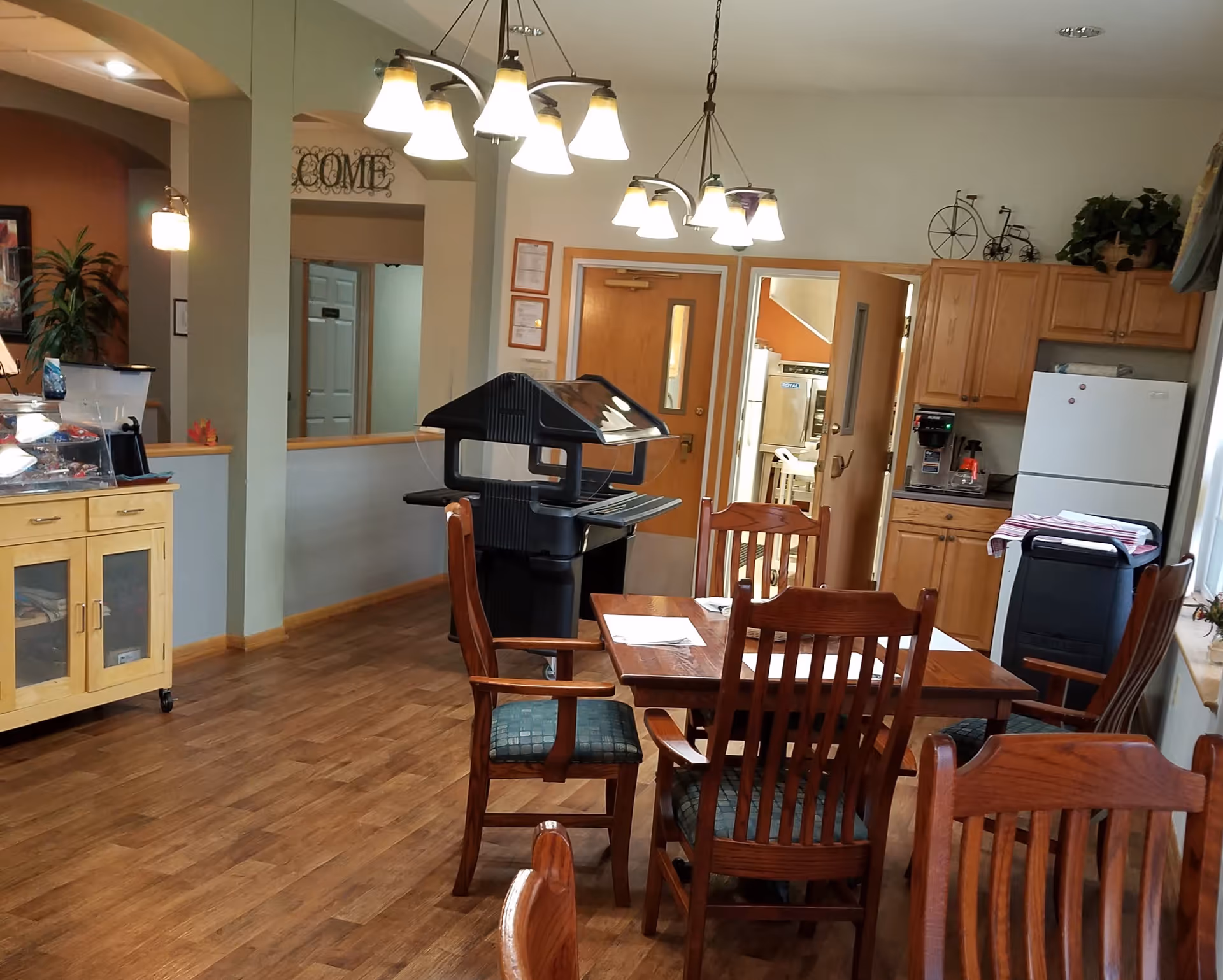 Interior view of a communal dining and kitchen area with wooden chairs and tables, a serving cart, a refrigerator, wooden cabinets, and overhead lighting fixtures. There is a doorway leading to another room and decorative items on top of the cabinets.