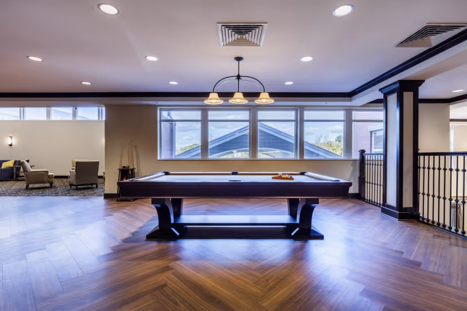 A spacious indoor recreational area featuring a pool table centered on a polished wooden floor with a chevron pattern. Behind the pool table are large windows showing the exterior roof and sky. To the left, there is a seating area with armchairs and a small table. The ceiling has recessed lighting and a hanging light fixture above the pool table. The room has a modern and clean design with dark trim accents.