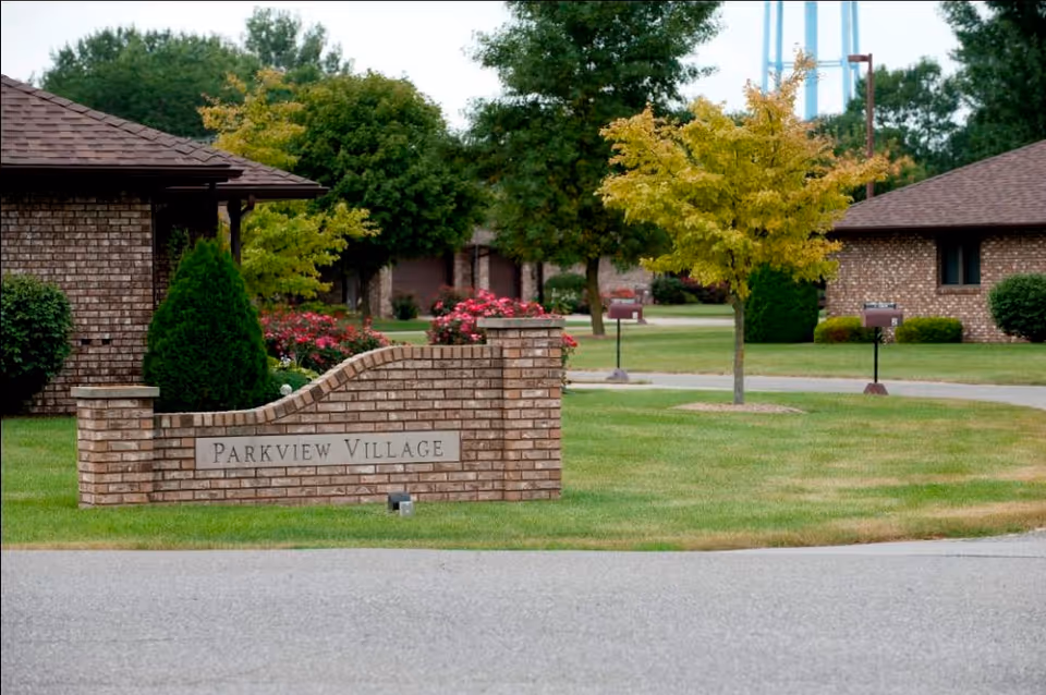 A brick entrance sign reading "Parkview Village" sits on a manicured lawn with trees, flowers, and single-story brick buildings in the background.