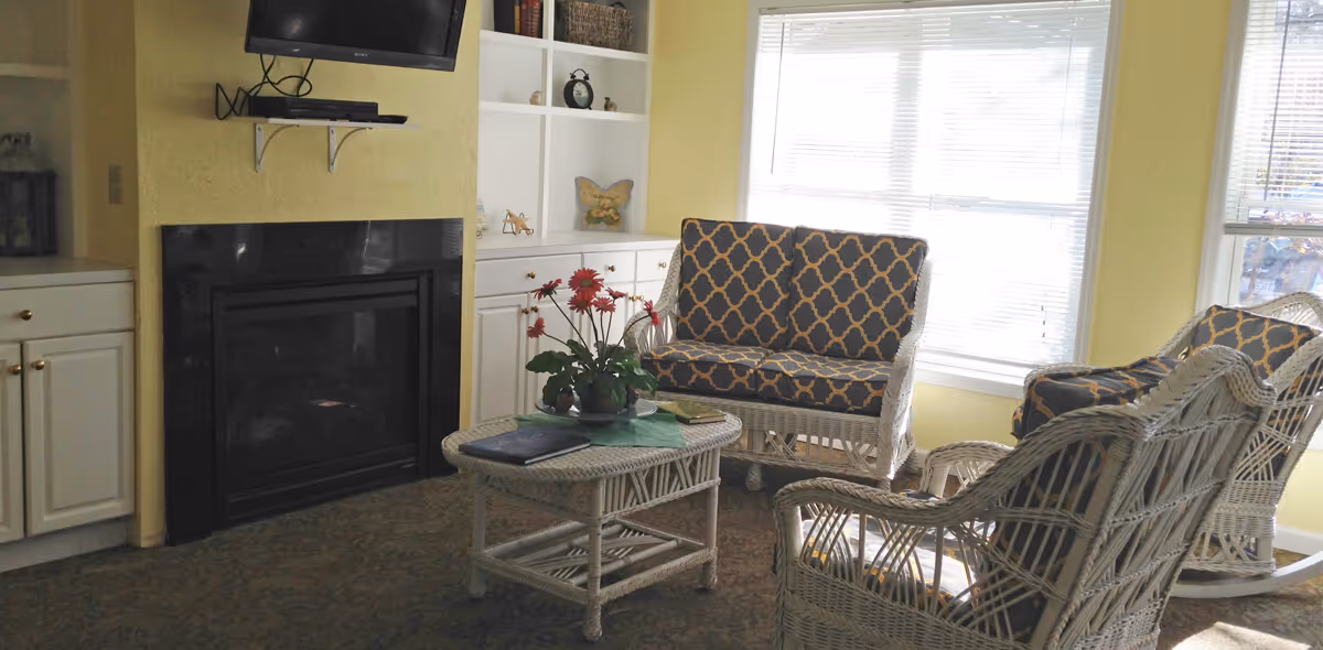 Sunlit sitting area with wicker chairs and a patterned loveseat around a coffee table, a fireplace beneath a wall-mounted TV, and built-in shelving.