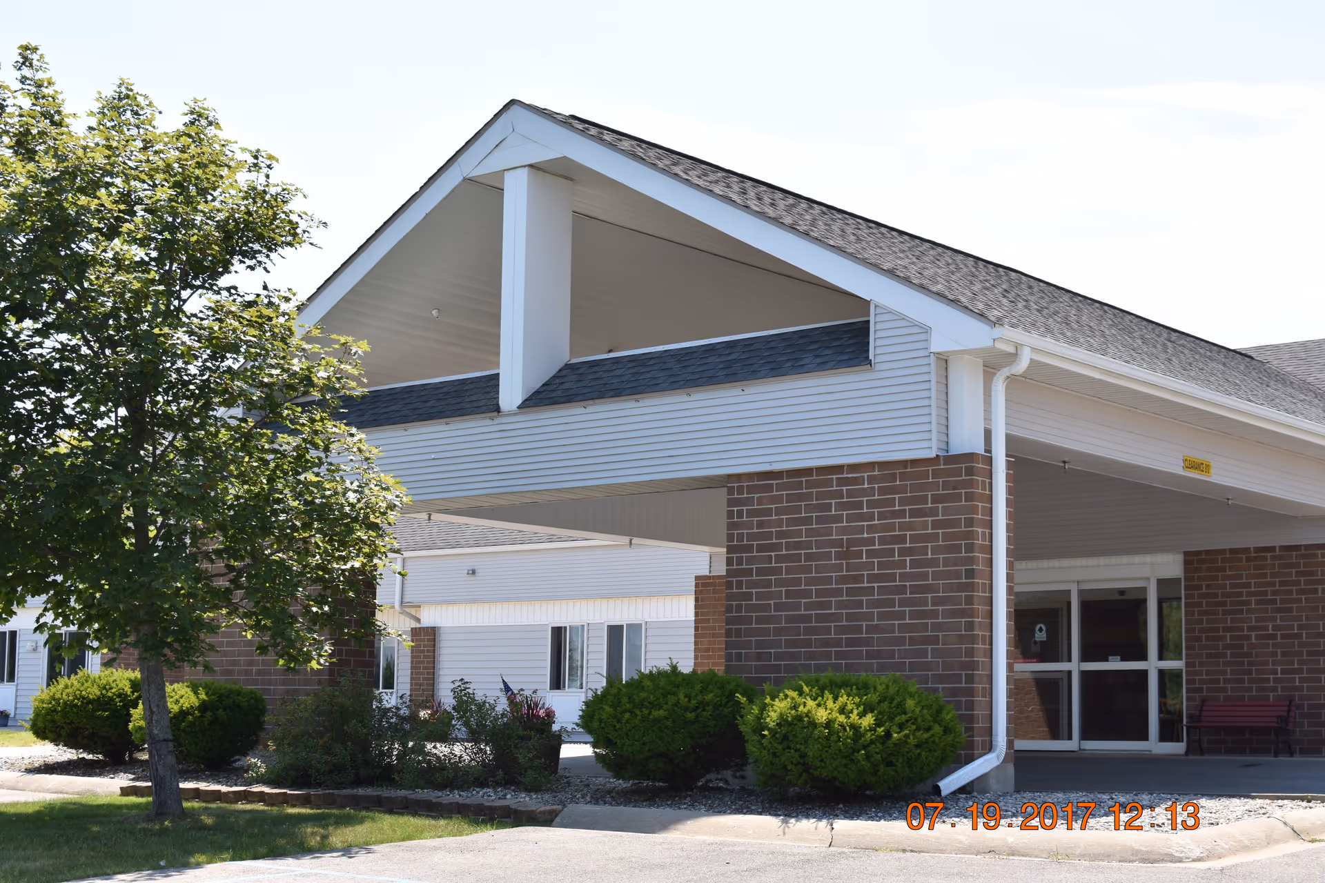 Entrance of a senior living facility with a covered drop-off area supported by brick pillars and white siding. There are green bushes and a tree near the entrance, and glass doors lead inside the building. The sky is clear and sunny.