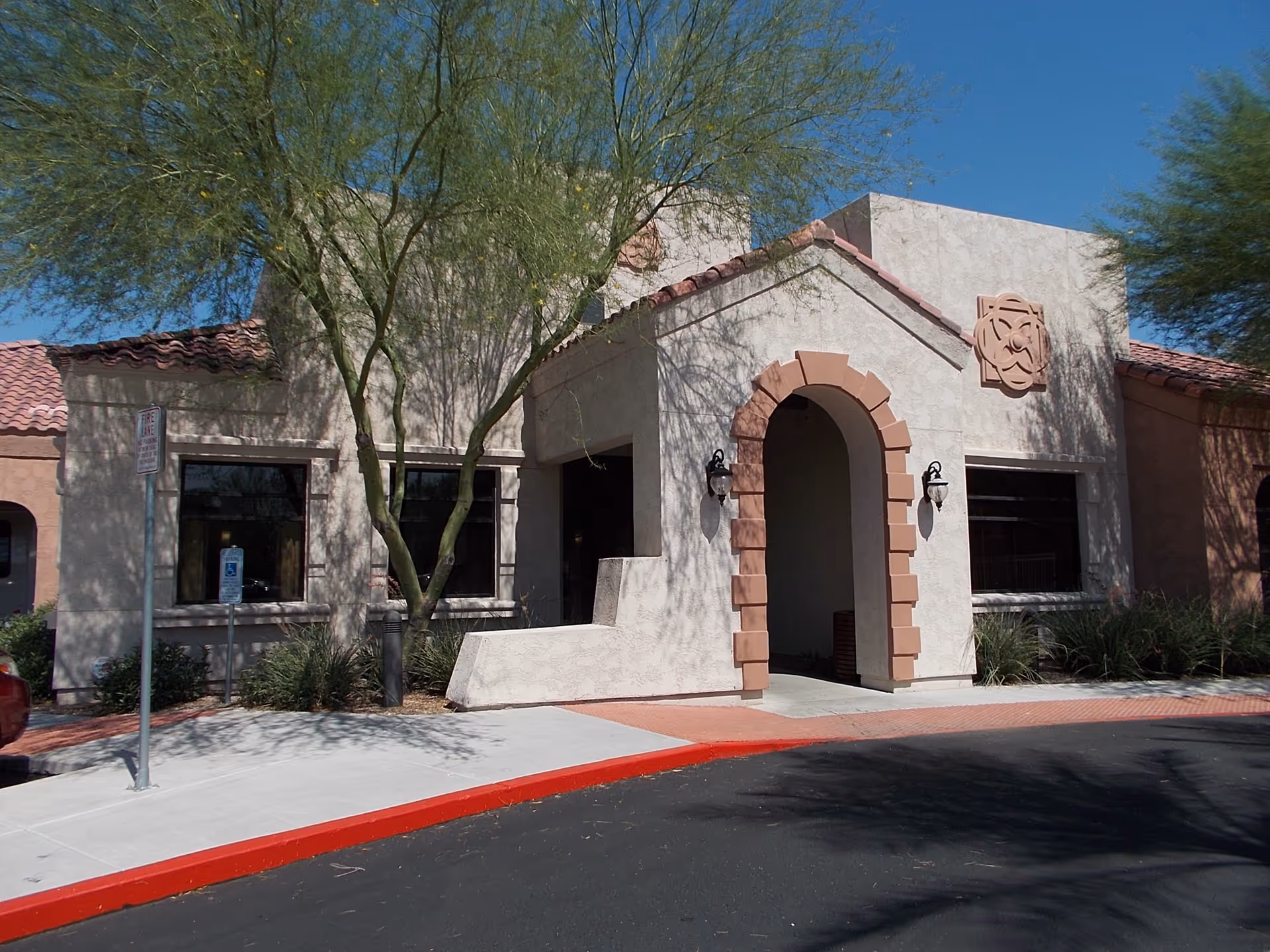 Front facade of a stucco building with a tiled roof, arched entryway, and desert landscaping.