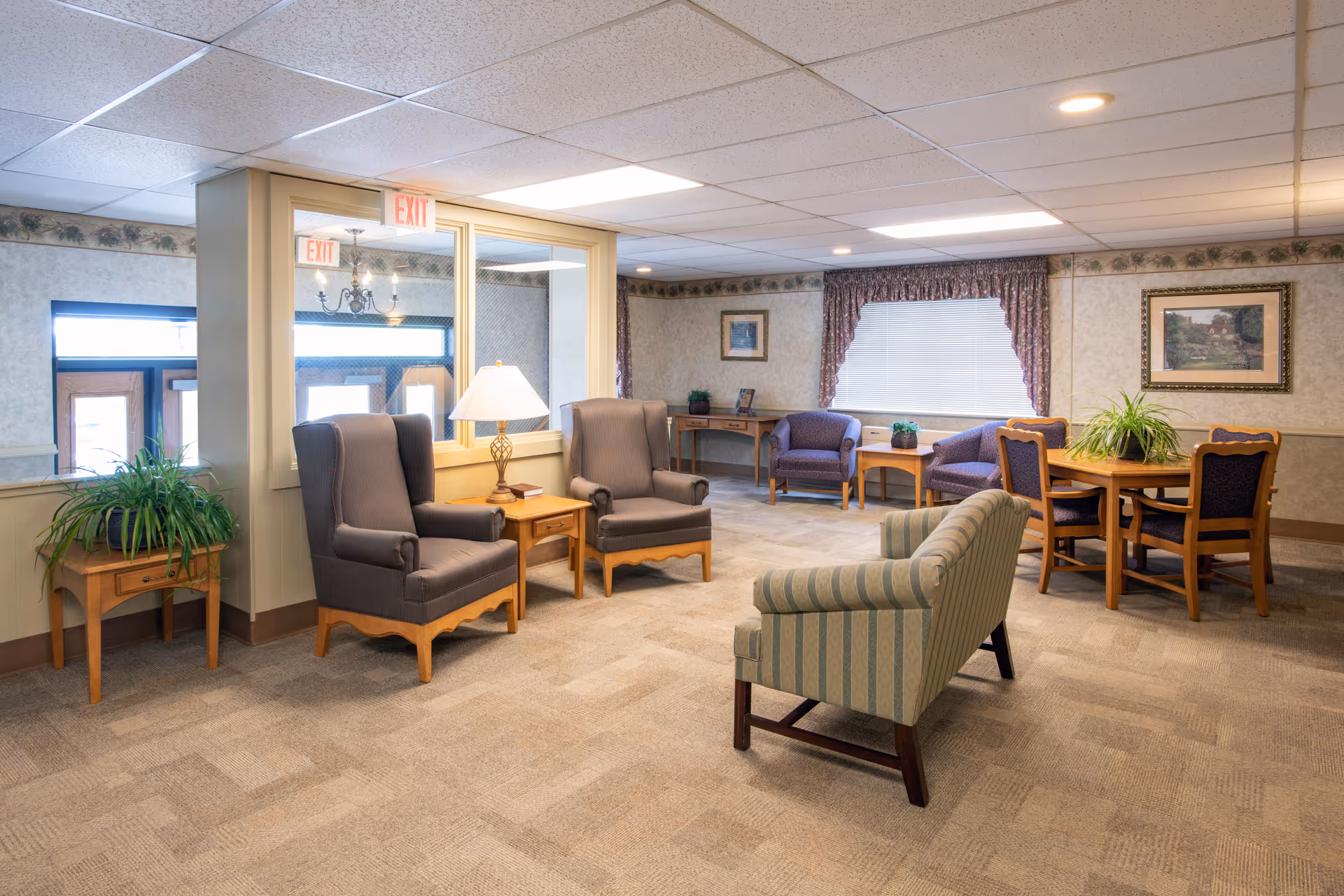 A well-lit common area in a senior living facility featuring several upholstered chairs and a small sofa arranged around wooden side tables and a round table with chairs. The room has carpeted floors, floral wallpaper border, framed artwork on the walls, and a window with blinds and curtains. There are also potted plants on the tables and a lamp on one of the side tables.