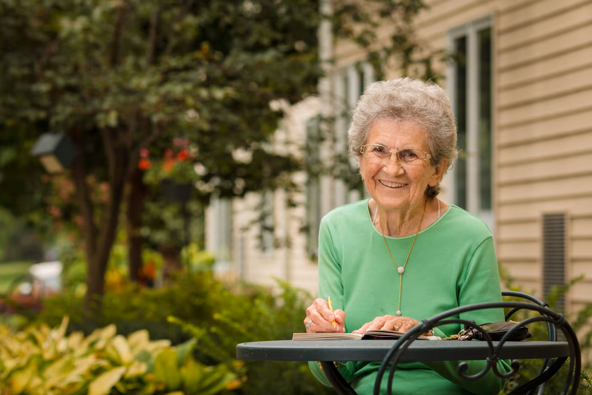 An elderly woman wearing glasses and a green top is sitting at a round outdoor table, smiling while writing in a notebook. The background shows greenery and the exterior wall of a building.
