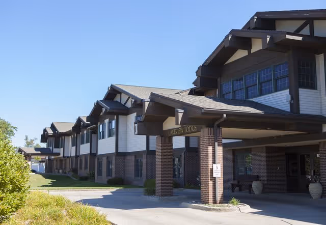 Exterior view of a senior living facility building named Lakeview Lodge with a covered entrance supported by brick columns, surrounded by greenery and a clear blue sky.