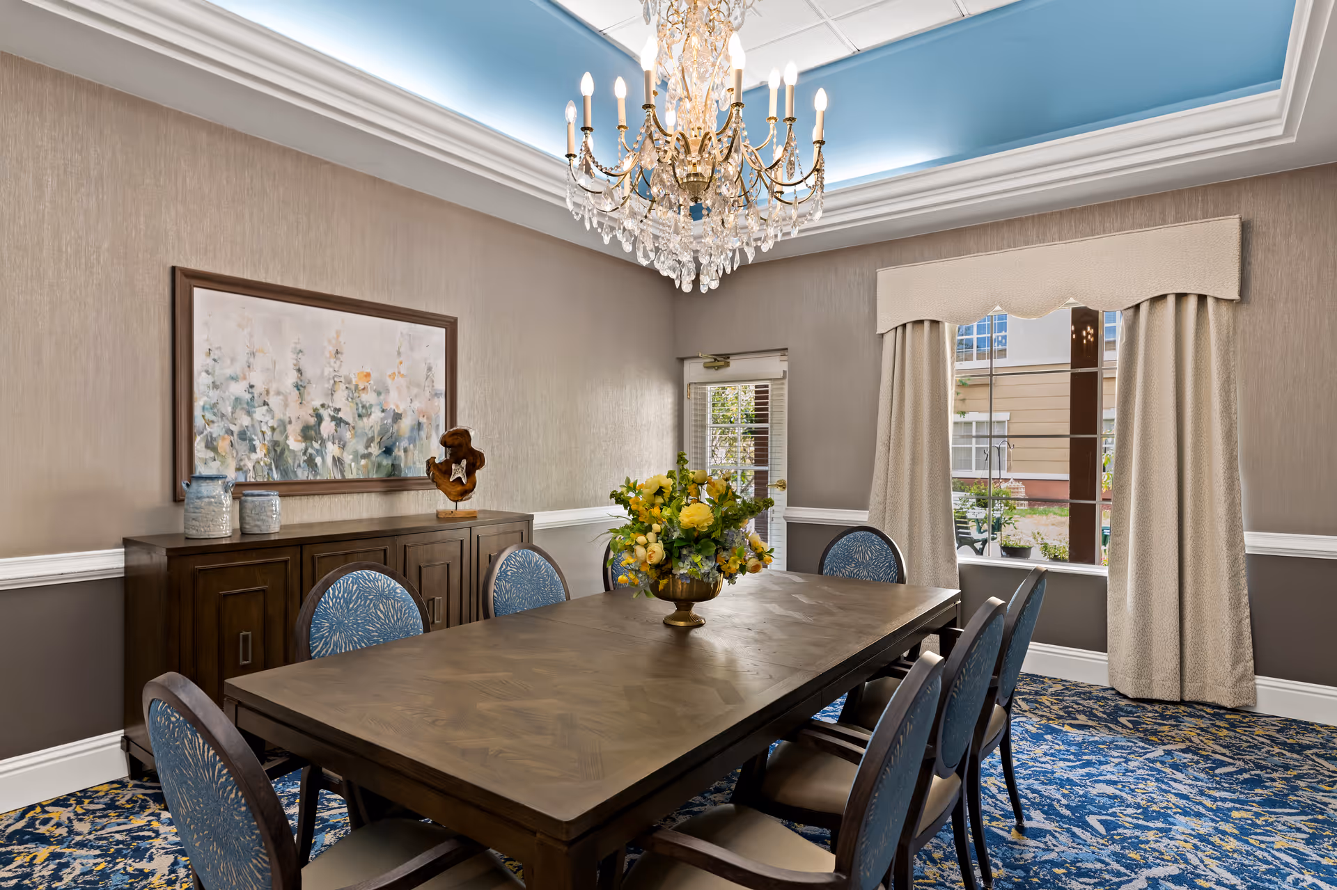 A formal dining room with a large wooden table surrounded by eight blue upholstered chairs. A decorative floral centerpiece sits on the table. The room features a crystal chandelier hanging from a recessed ceiling with blue accent lighting. There is a sideboard against one wall with decorative items and a large framed floral painting above it. A window with beige curtains allows natural light into the room.