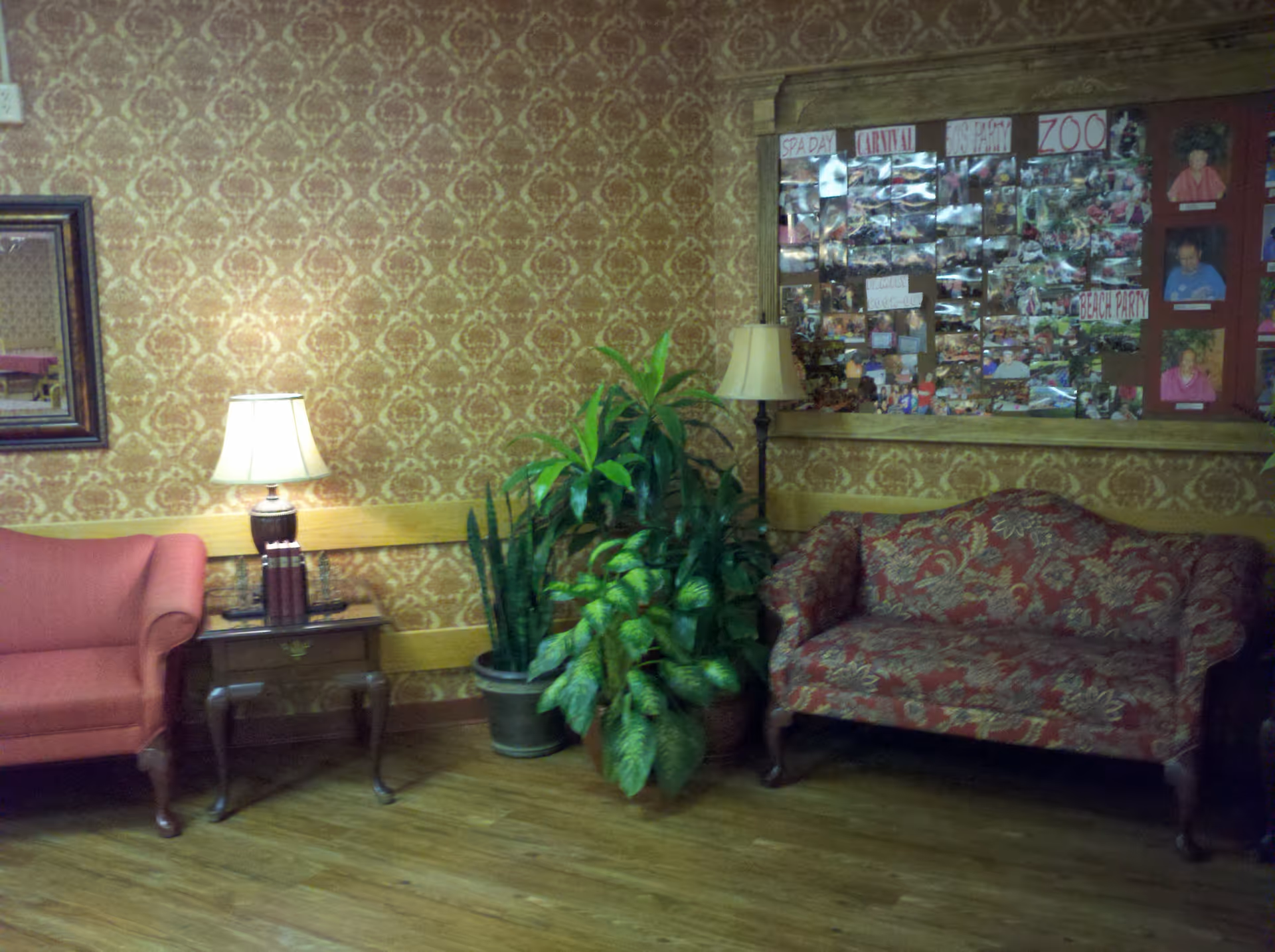 Lobby-style seating area with two upholstered sofas, a side table and lamp, potted plants, and a photo bulletin board on patterned wallpaper.