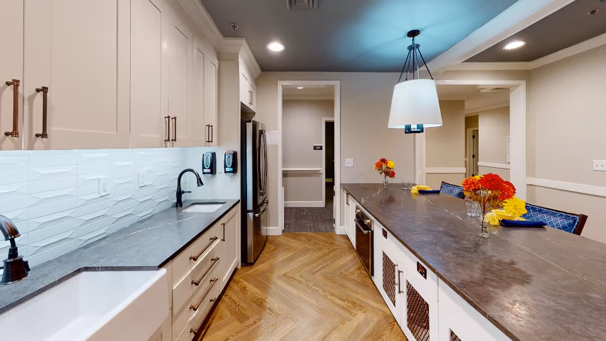 A modern kitchen with white cabinetry, a dark countertop, and a white farmhouse sink. The backsplash features a textured white tile design. There is a stainless steel refrigerator and two wall-mounted soap dispensers. A large island with a dark countertop has three vases with red and yellow flowers, blue napkins, and chairs with blue cushions. The floor has a herringbone wood pattern, and a pendant light hangs above the island.