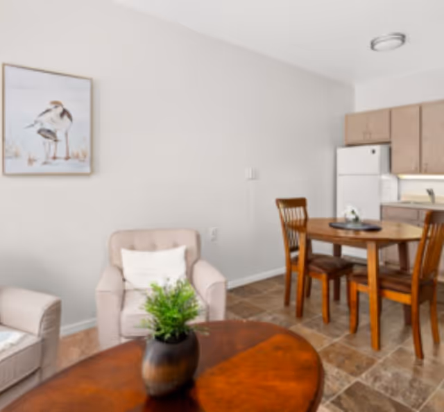 A cozy interior space featuring a small dining area with a round wooden table and four chairs, a kitchenette with wooden cabinets and a white refrigerator, and a seating area with a beige armchair and a matching loveseat. A framed picture of birds hangs on the wall, and a small potted plant sits on a wooden coffee table in the foreground.