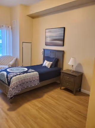 A bedroom with a single bed featuring a dark blue bedspread with patterned designs. There is a wooden nightstand with a white lamp on it next to the bed. A framed picture of a serene landscape hangs on the beige wall above the bed. To the left, there is a window with white curtains and a cushioned chair with a geometric pattern.