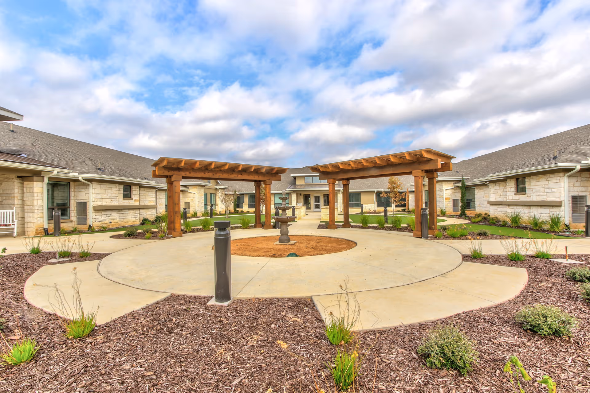Outdoor courtyard area at Long Creek facility featuring two wooden pergolas, a central circular concrete pathway with a small fountain in the middle, surrounded by landscaped garden beds with mulch and plants, and single-story stone buildings with gray roofs in the background under a partly cloudy sky.