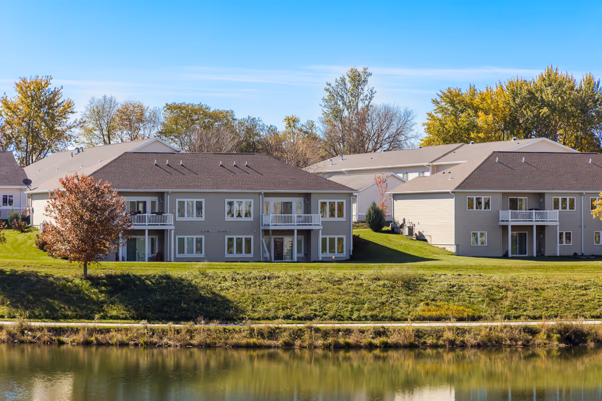 View of a senior living facility named Glenwood Place showing multiple two-story residential buildings with balconies and large windows, surrounded by green lawns and trees, with a calm body of water in the foreground reflecting the buildings and trees under a clear blue sky.
