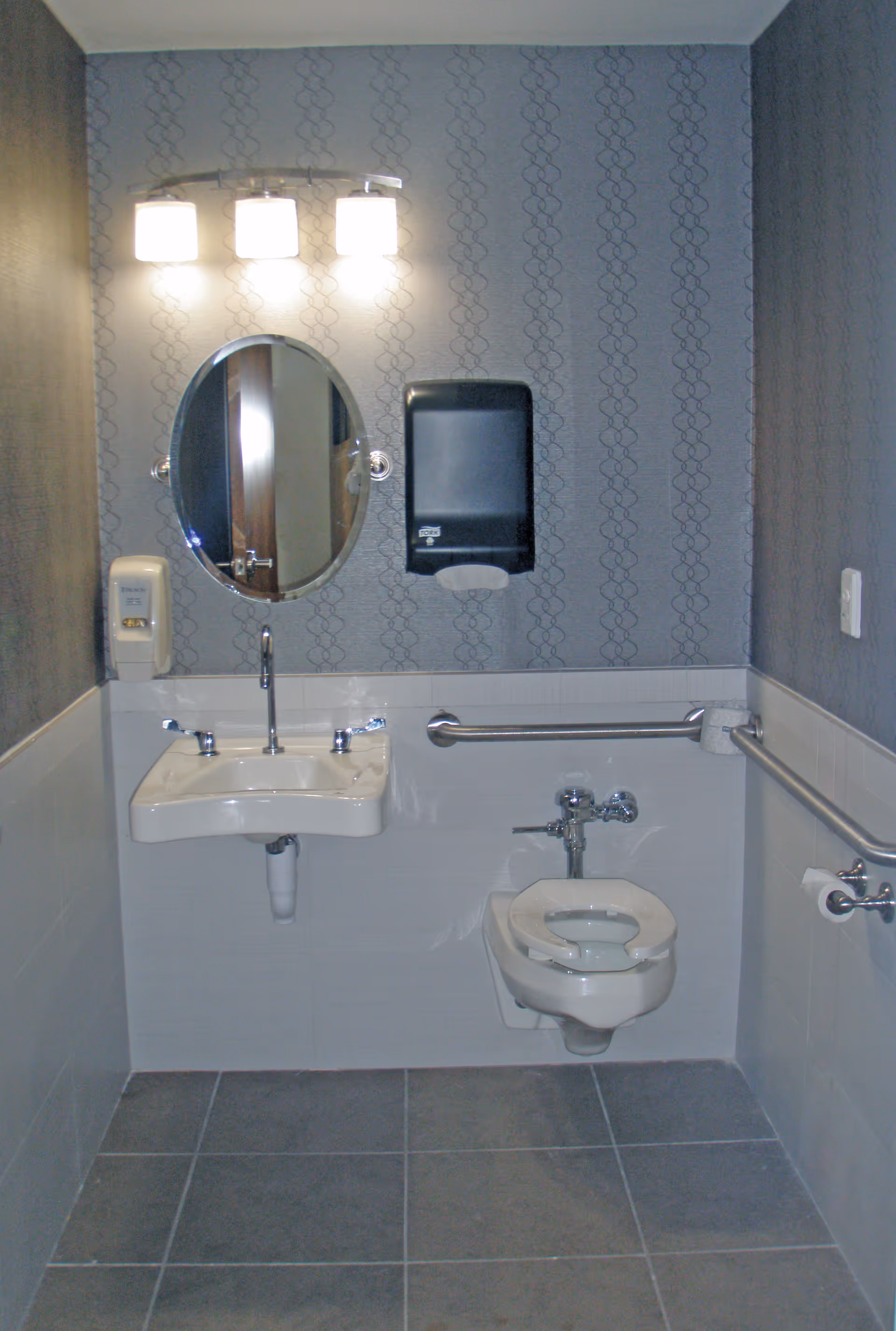 A small, accessible bathroom with a wall-mounted sink and faucet, an oval mirror above the sink, a soap dispenser on the left wall, a paper towel dispenser above the sink, a toilet with a raised seat and grab bars on both sides, and a toilet paper holder on the right wall. The walls have a patterned wallpaper above white tiles, and the floor is tiled in gray.