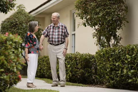 An elderly man and woman holding hands on a sidewalk beside a single-story building with shrubs.