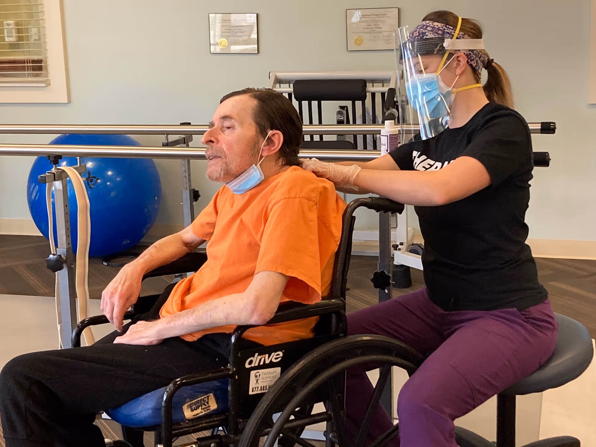 A man in an orange shirt sitting in a wheelchair receiving a shoulder massage from a caregiver wearing a face mask, face shield, gloves, and a headband in a therapy or rehabilitation room with exercise equipment in the background.