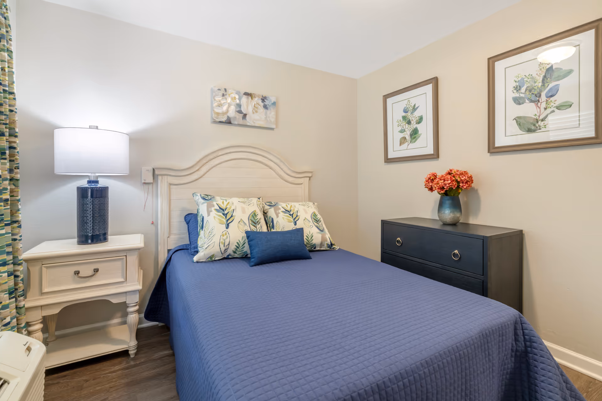 A cozy bedroom with a bed covered in a blue quilt and decorative pillows with leaf patterns. There is a white nightstand with a blue lamp on the left side of the bed, and a dark dresser with a vase of red flowers on top on the right side. The walls are beige and decorated with floral artwork.