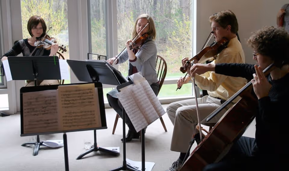 Four musicians seated indoors playing string instruments including violins and a cello, with music stands and sheet music in front of them, and large windows showing greenery outside.