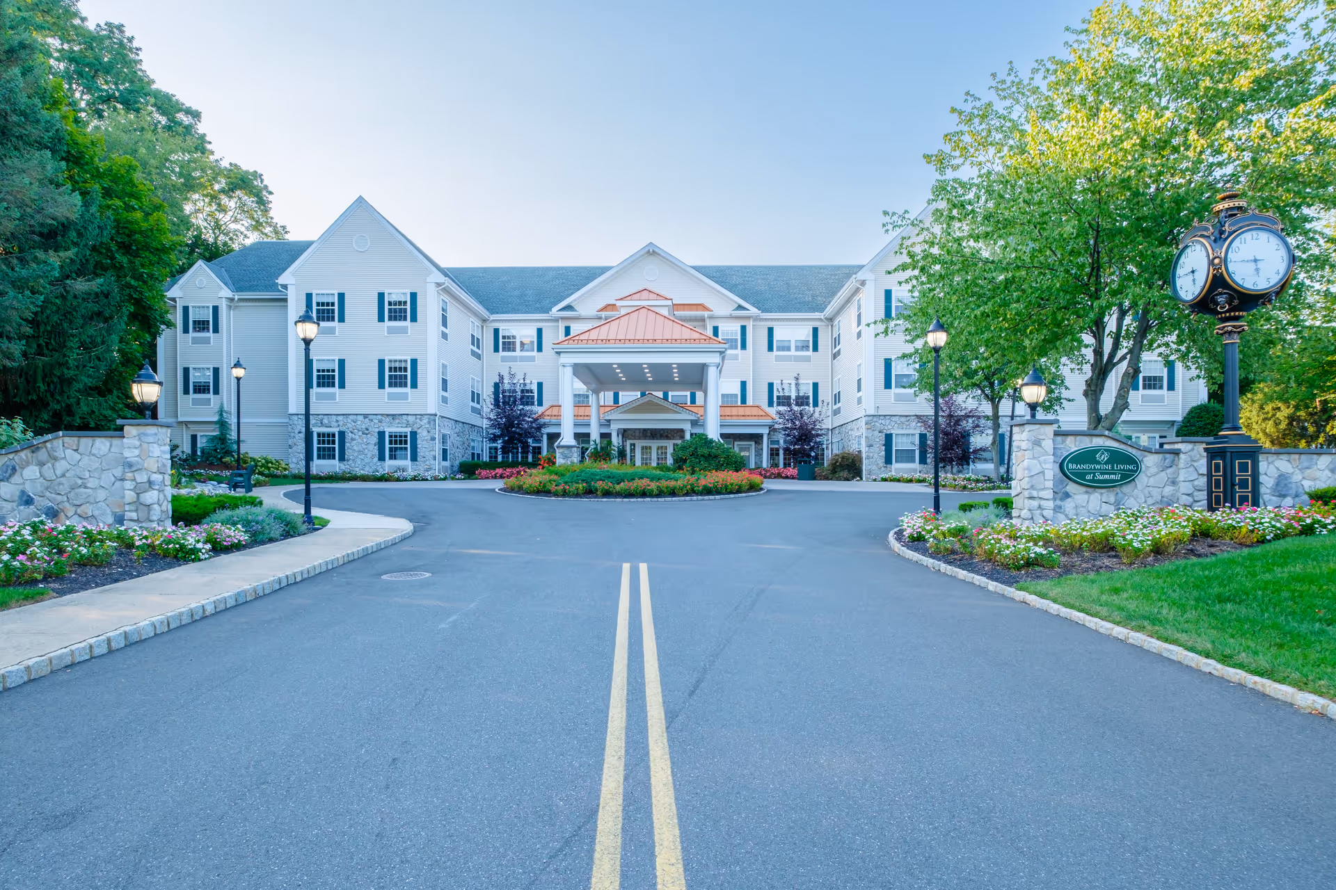 Front exterior view of Brandywine Summit by Monarch, a multi-story senior living facility with a circular driveway, landscaped flower beds, lamp posts, and a decorative clock on the right side.