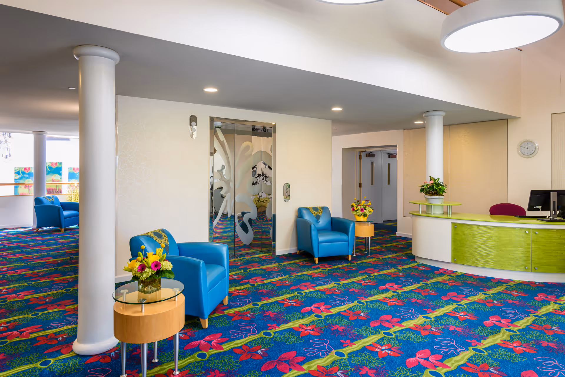 A bright and colorful lobby area with a vibrant floral carpet, two blue armchairs each accompanied by a small round table with flower arrangements, a green reception desk with a computer, and an elevator with decorative glass doors. The space is well-lit with ceiling lights and has white columns and walls.