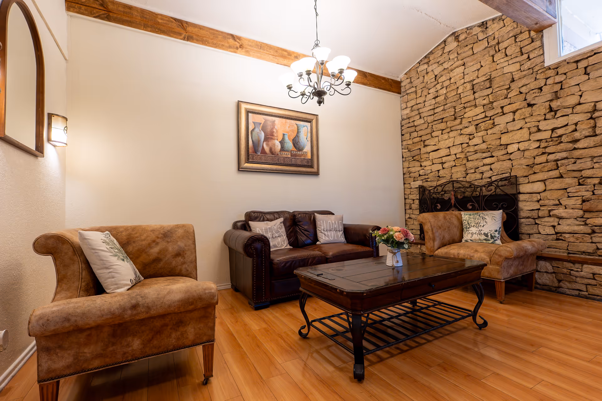 A cozy living room with two brown leather armchairs and a dark brown leather loveseat arranged around a wooden coffee table with a flower vase. The room features a stone accent wall with a decorative fireplace screen, wooden flooring, a chandelier hanging from the ceiling, and a framed painting of vases on the wall.