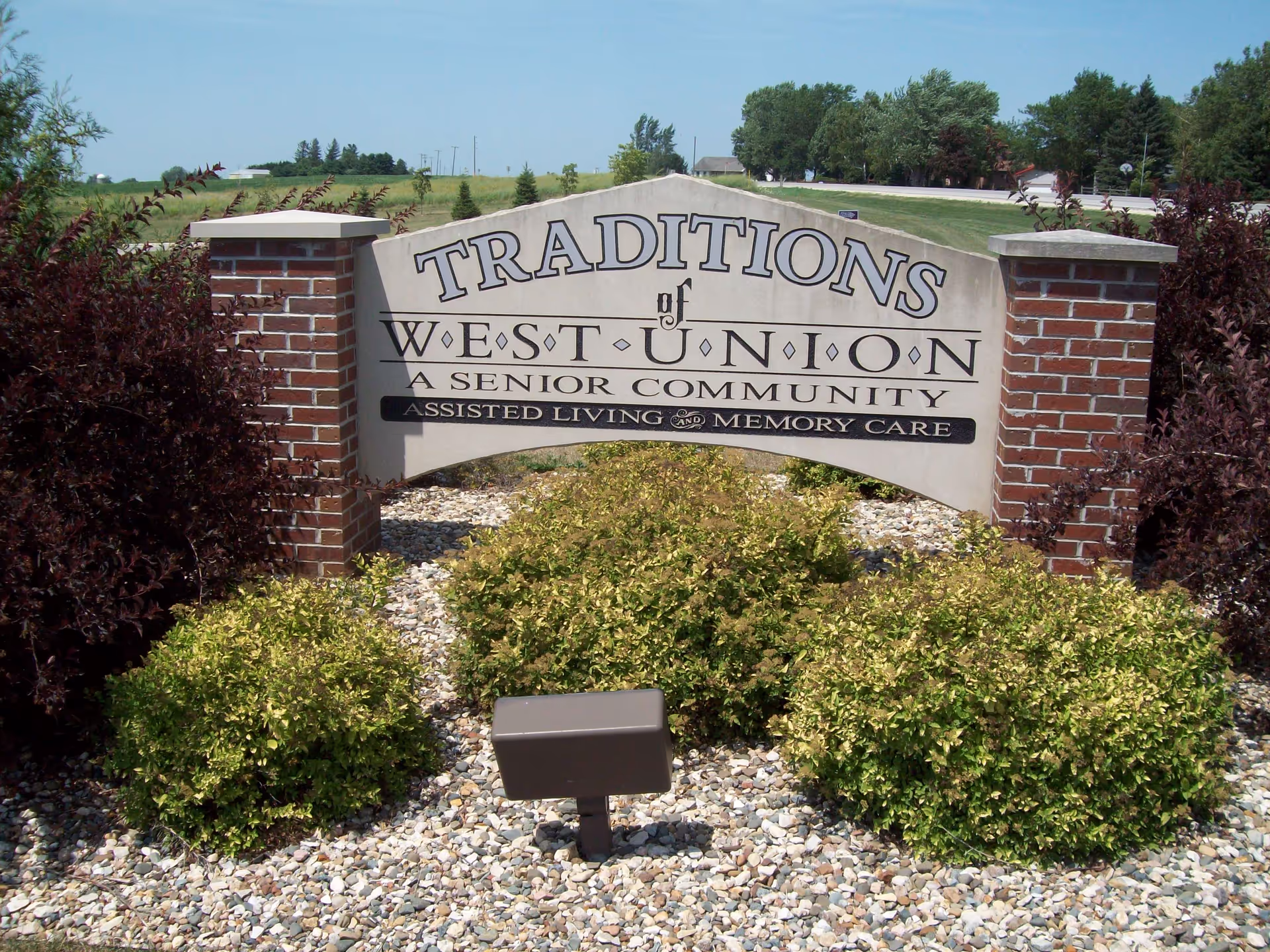 Stone and brick entrance sign reading "Traditions of West Union, a senior community — assisted living and memory care" set among shrubs and gravel.