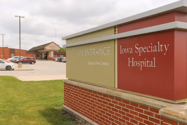Exterior view of Iowa Specialty Hospital with a large sign in the foreground indicating the main entrance and tobacco free campus. The hospital building and parking lot with several cars are visible in the background under a cloudy sky.