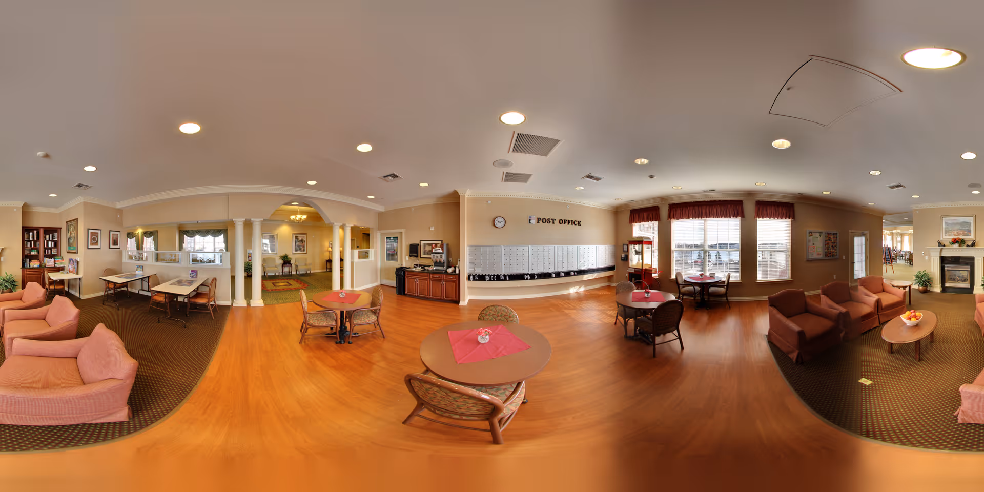 A spacious senior living common area with multiple seating arrangements including pink and brown armchairs and small round tables with red tablecloths. There is a post office section with mailboxes on the wall, large windows with red valances, and a fireplace on the right side. The room has warm lighting, wooden floors, and beige walls with framed artwork and plants.