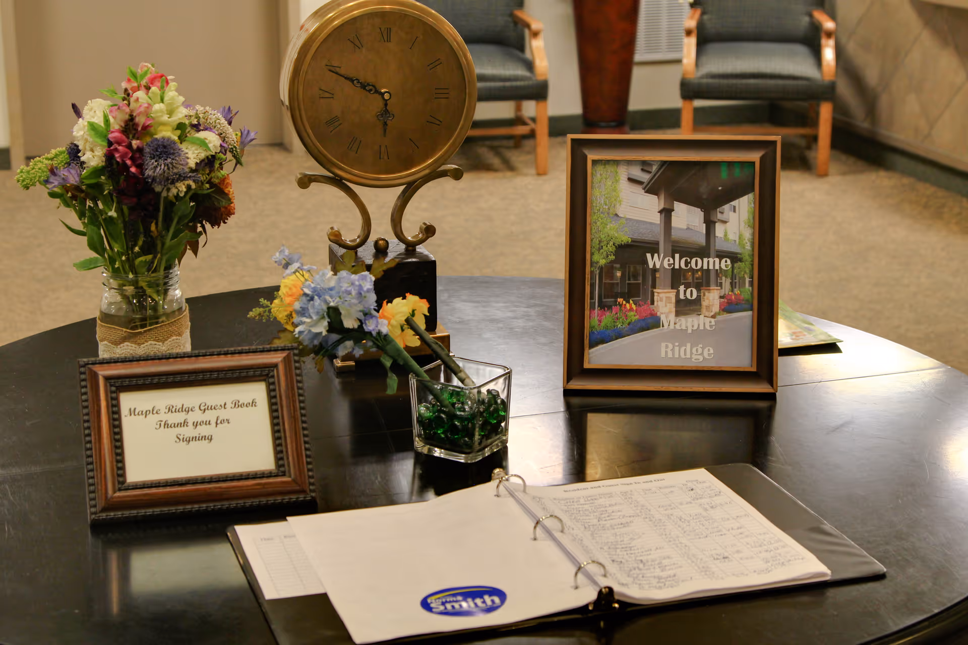 A round reception table with a guest book binder, framed 'Welcome to Maple Ridge' sign, flowers, and a decorative clock in a lobby.