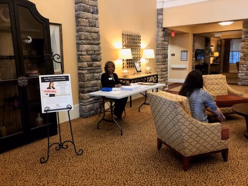 A woman sitting behind a table with papers in a warmly lit lobby area with stone pillars and patterned carpet. Another person is seated on a patterned armchair facing away. A sign on a stand near the table indicates hiring information for Landon Ridge Sugar Land Independent Living.