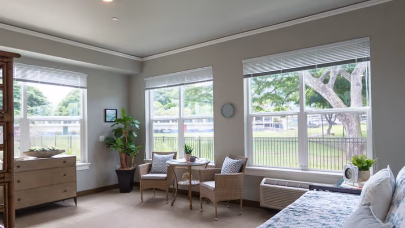 Sunlit senior living bedroom with large windows, two wicker chairs and a small table, a dresser, plants, and a bed.
