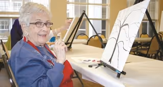An elderly woman with white hair and glasses is sitting at a table in a well-lit room, smiling while painting on a canvas set on an easel. The canvas shows the outline of a tree. Other easels and chairs are visible in the background.