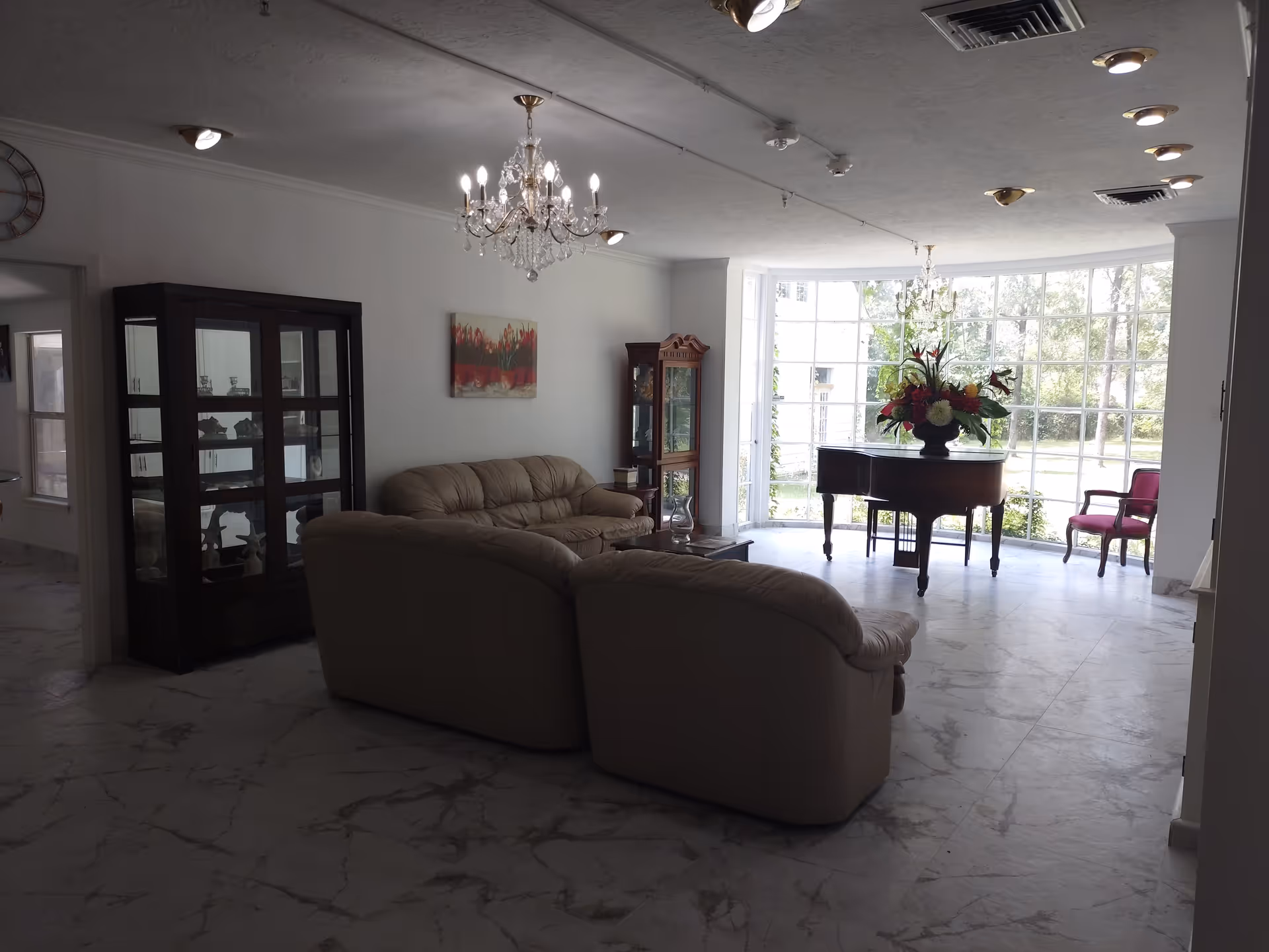 A bright living room with large floor-to-ceiling windows letting in natural light. The room features a beige leather sofa set, a glass display cabinet, a wooden grandfather clock, a chandelier, a grand piano with a floral arrangement on top, and a single red upholstered chair near the window. The floor is covered with white marble tiles with gray veining.