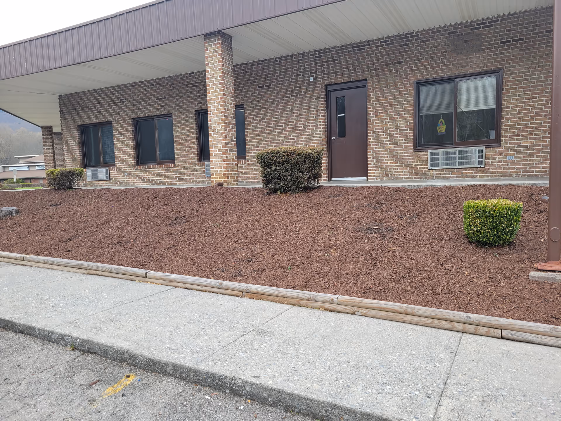 Brick exterior of a single-story building with a covered entrance, several windows, a door, and a mulch bed with small shrubs.