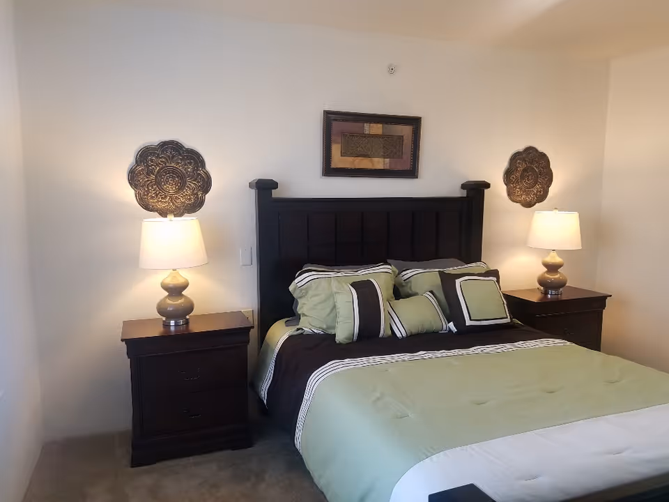 A neatly made bed with a dark wooden headboard, green and brown bedding, and multiple pillows. On each side of the bed are matching dark wooden nightstands with table lamps. Decorative wall art is hung above the headboard and on either side of the bed. The room has light-colored walls and carpeted flooring.