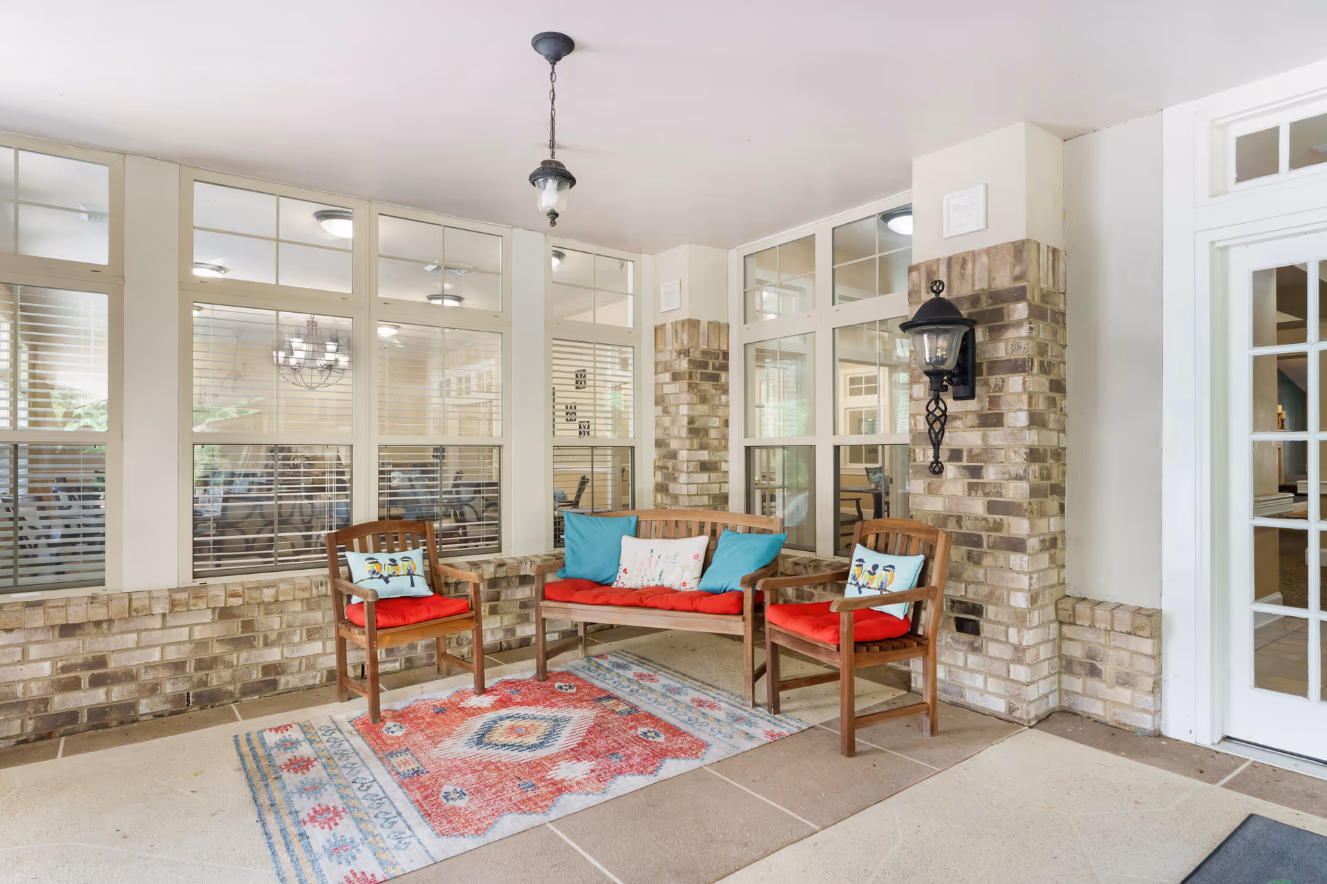 Covered outdoor seating area with two wooden chairs and a wooden bench, all with red cushions and decorative pillows. The area features a colorful patterned rug on the floor, brick pillars, large windows with white frames, and a hanging light fixture above.