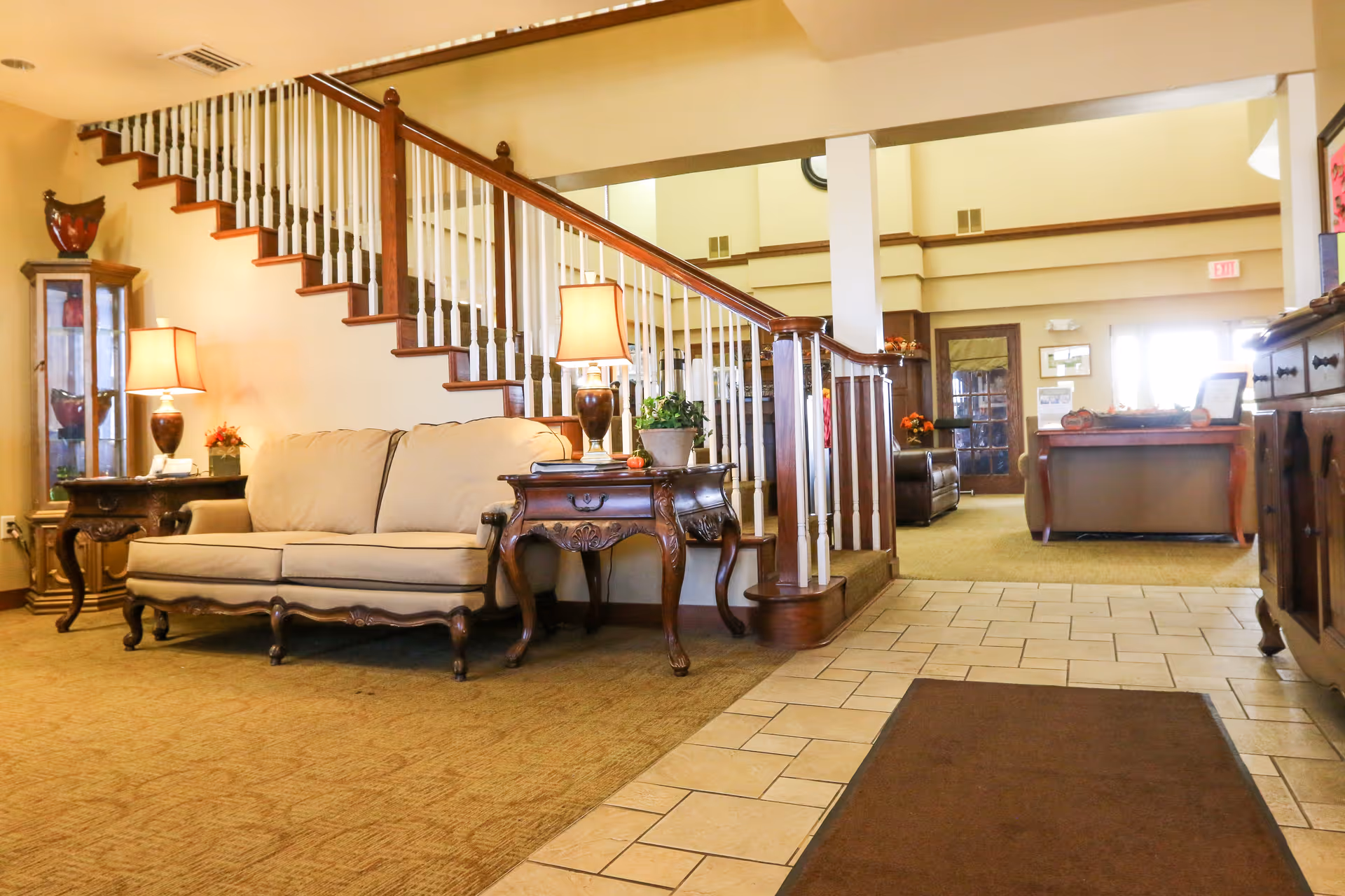A warm lobby with a sofa, side tables and lamps in front of a wooden staircase leading to an upper level.