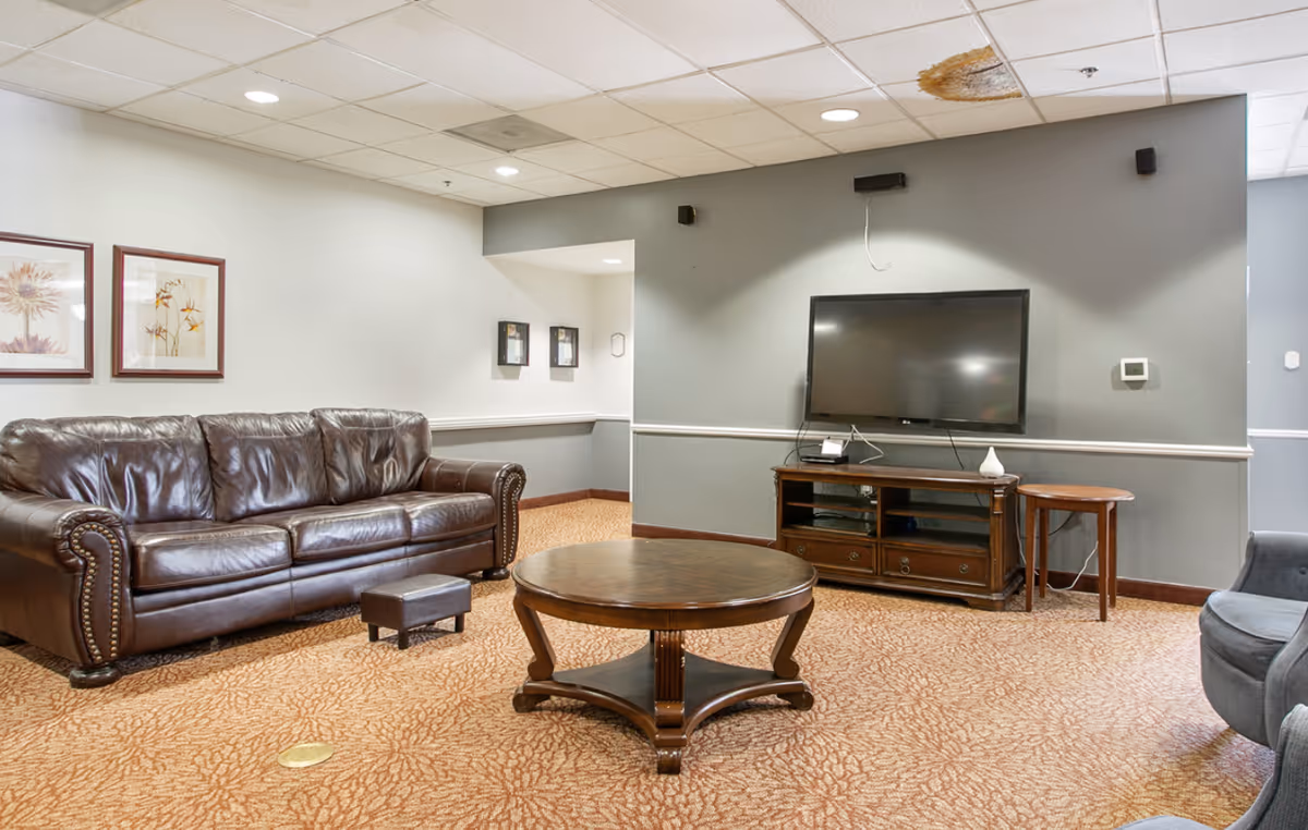 A senior living lounge with a leather sofa, round wooden coffee table, TV on a media console, and patterned carpeting.