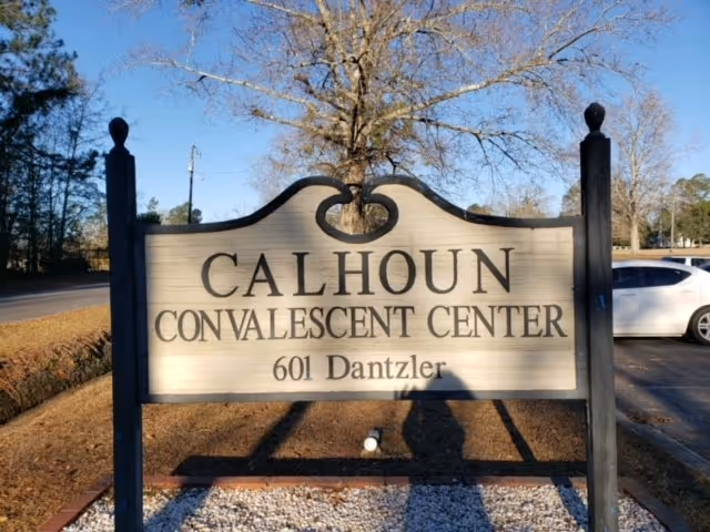 Outdoor wooden sign for Calhoun Convalescent Center at 601 Dantzler, with trees and a parked white car in the background under a clear blue sky.
