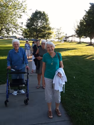 A group of elderly women walking along a paved path in a park-like outdoor setting with green grass and trees on either side. One woman in front is smiling and holding a white jacket, while another uses a walker. The sun is shining brightly.